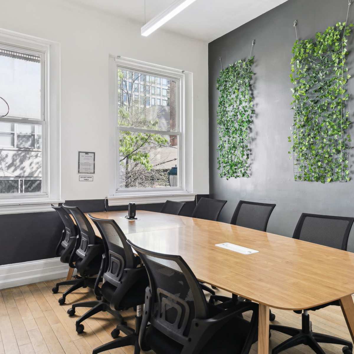 A conference room features a long wooden table surrounded by black chairs, with green wall decor and large windows.