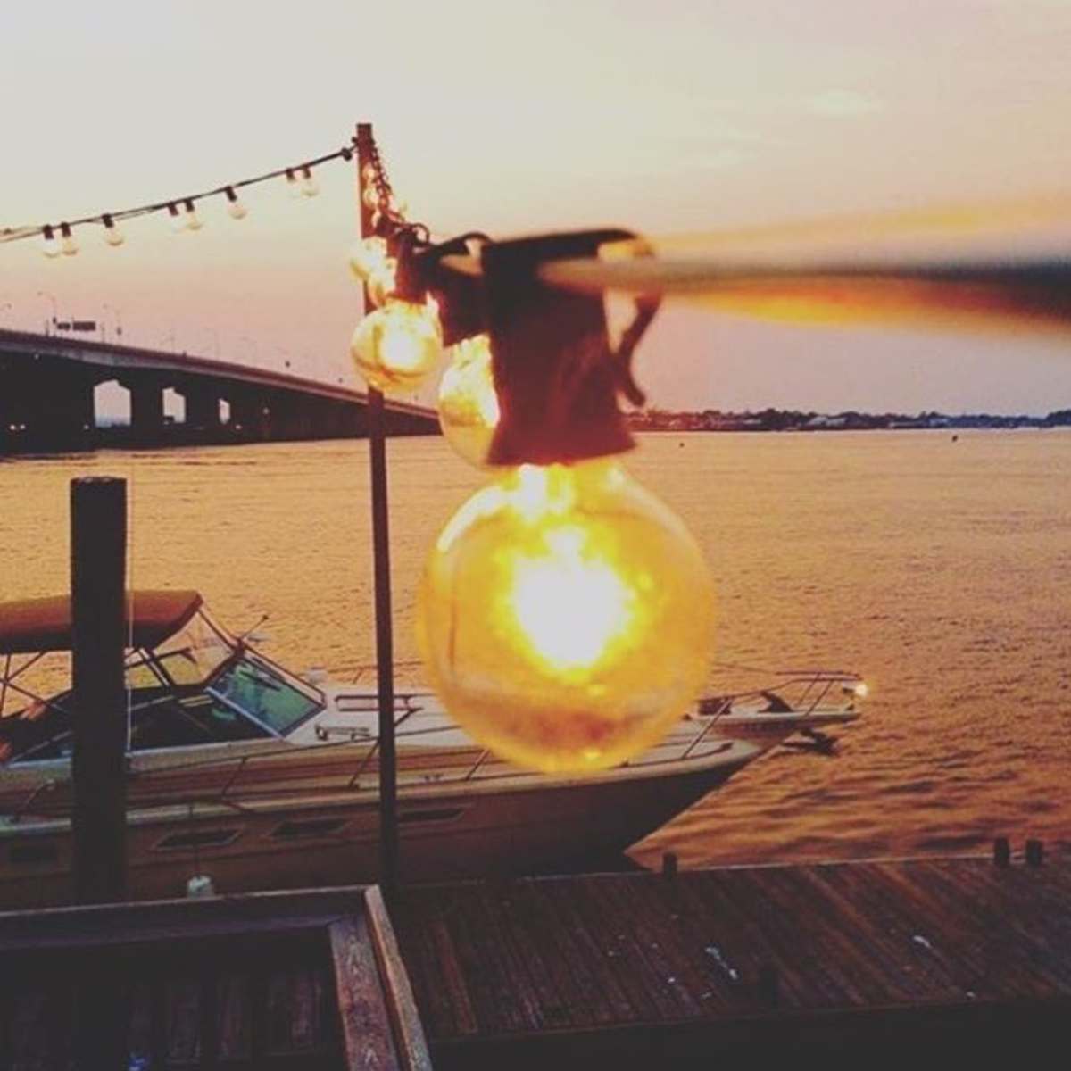 A string of illuminated bulbs hanging above a boat docked near the water as the sun sets in the background.