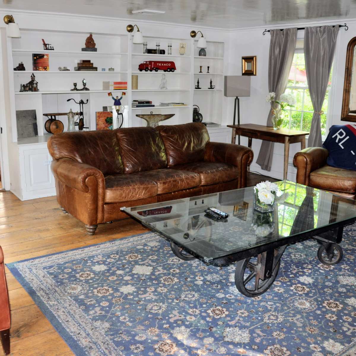 A living room featuring a leather sofa, a glass coffee table on a patterned rug, and bookshelves filled with various decorative items.