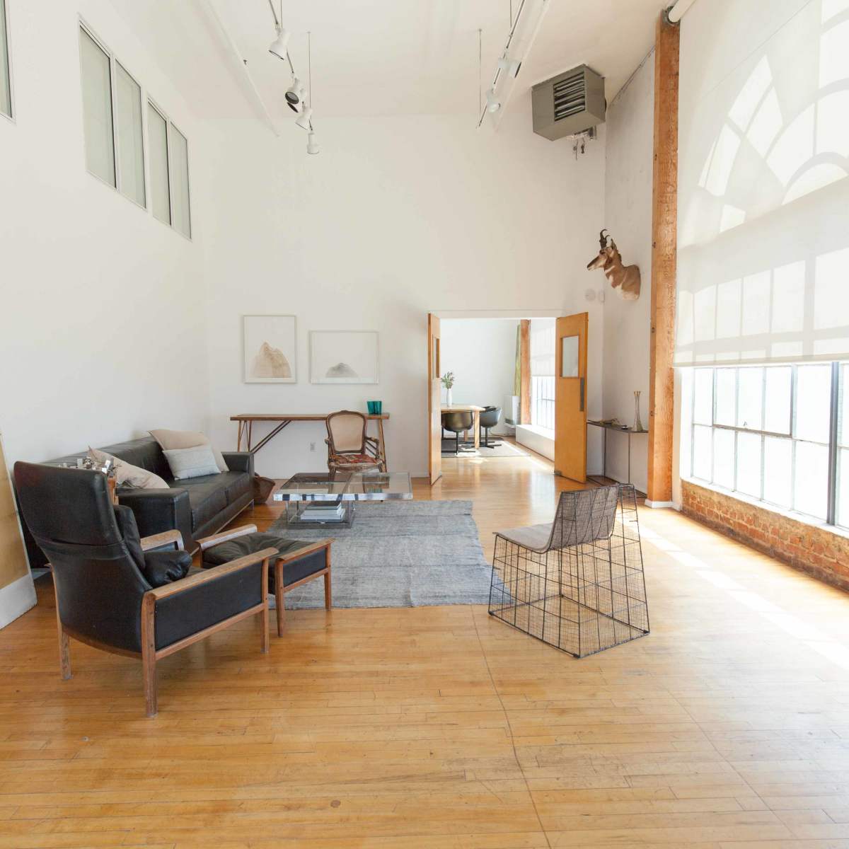 A spacious, well-lit loft-style room with hardwood floors and minimalistic furniture, including a sofa, a wire chair, and large windows.