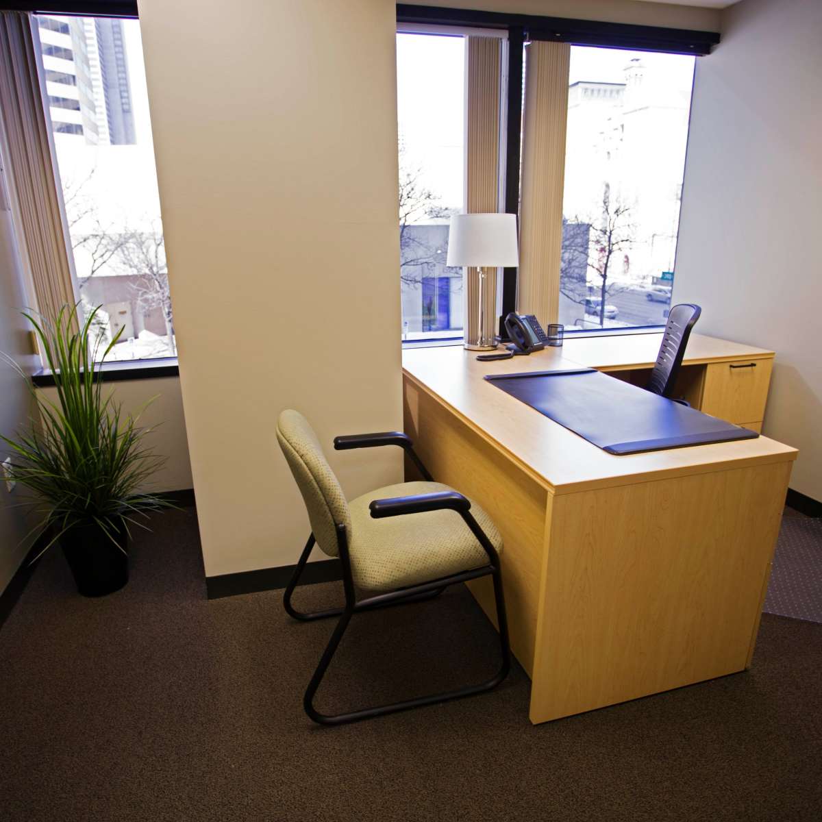 An office space features a wooden desk with a phone, a lamp, and two chairs near large windows.