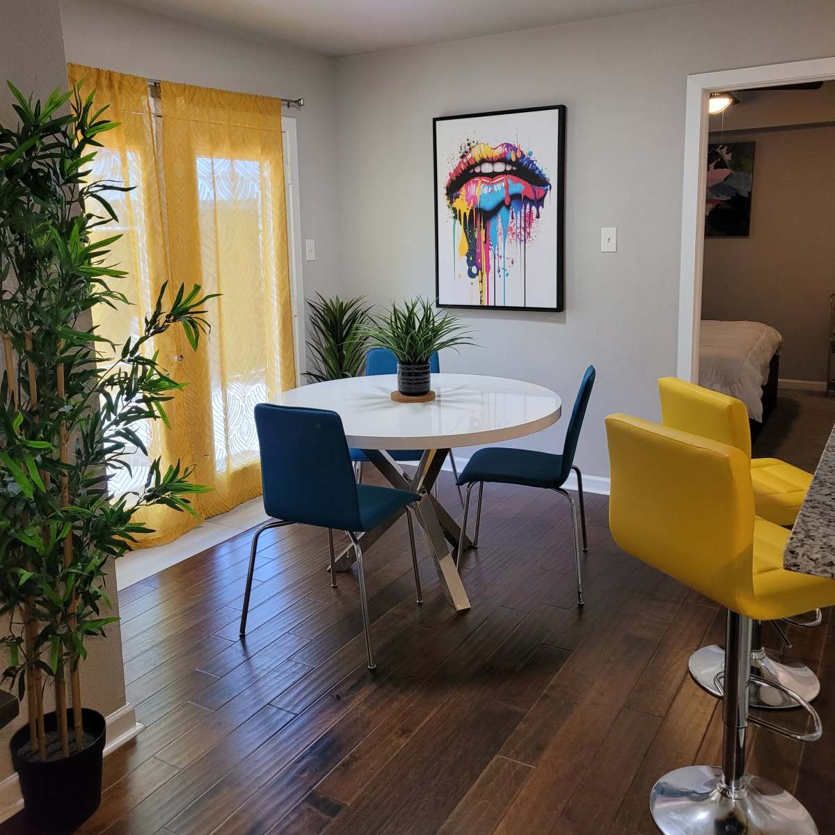 A dining area features a round table surrounded by four chairs, with vibrant yellow and blue accents, and a large art piece on the wall.