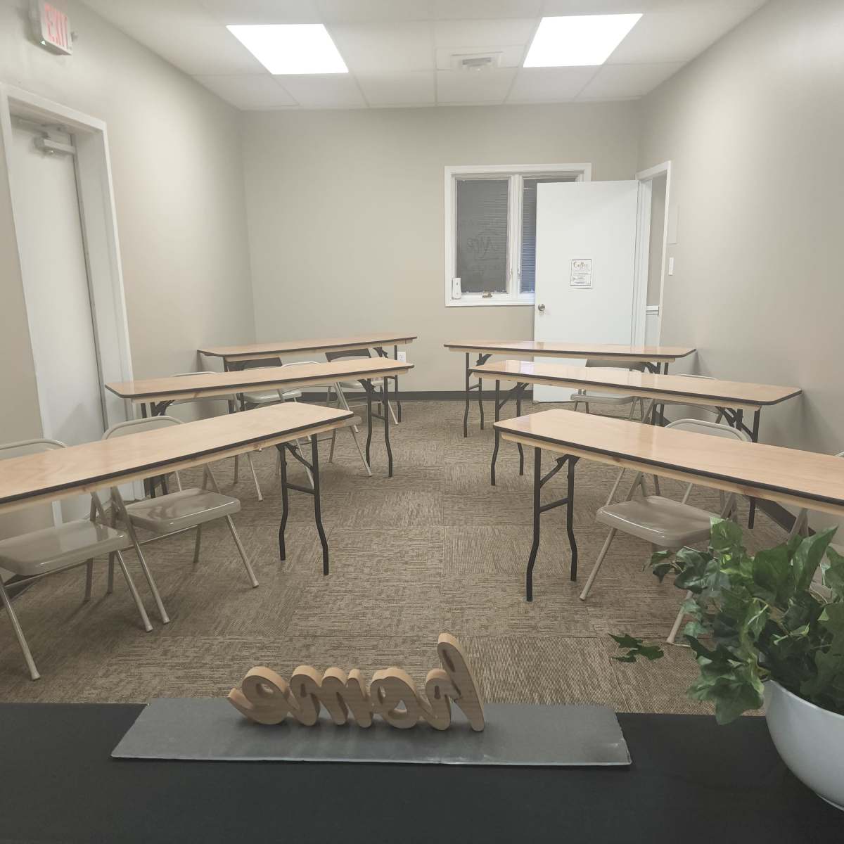 A small, vacant classroom with several tables arranged in a U-shape and a plant on a desk at the front.