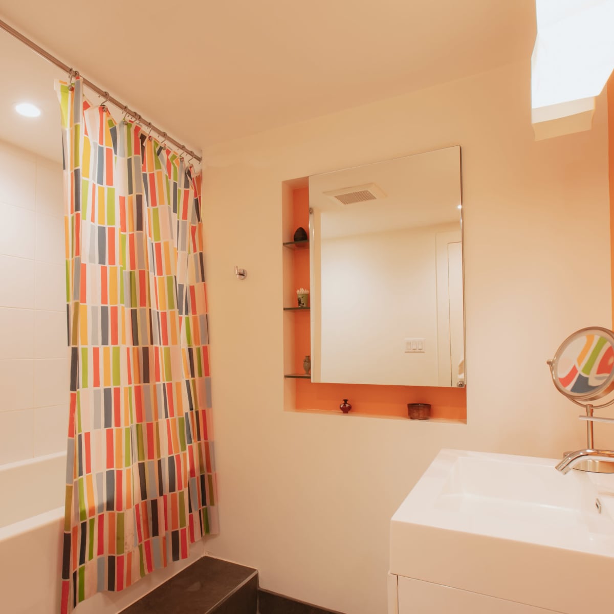 The bathroom features a shower with a colorful striped curtain, a rectangular mirror above a sink, and a wall-mounted shelf.
