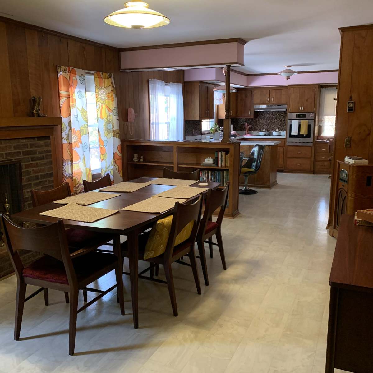 A dining area with a wooden table and chairs, leading into a kitchen with wooden cabinetry and floral curtains.