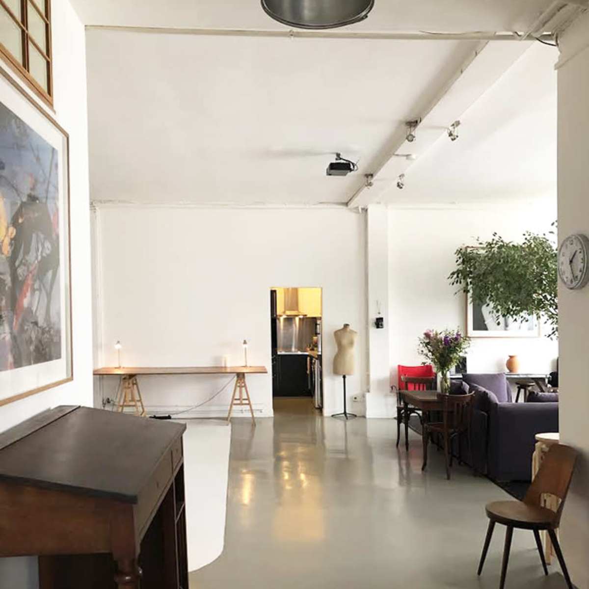 A spacious, well-lit interior of a room with a wooden desk, a chair, and a view into a kitchen area.