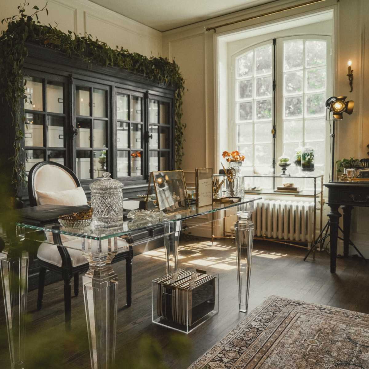 A stylish office space featuring a glass desk, a black cabinet with shelves, and large windows allowing natural light.