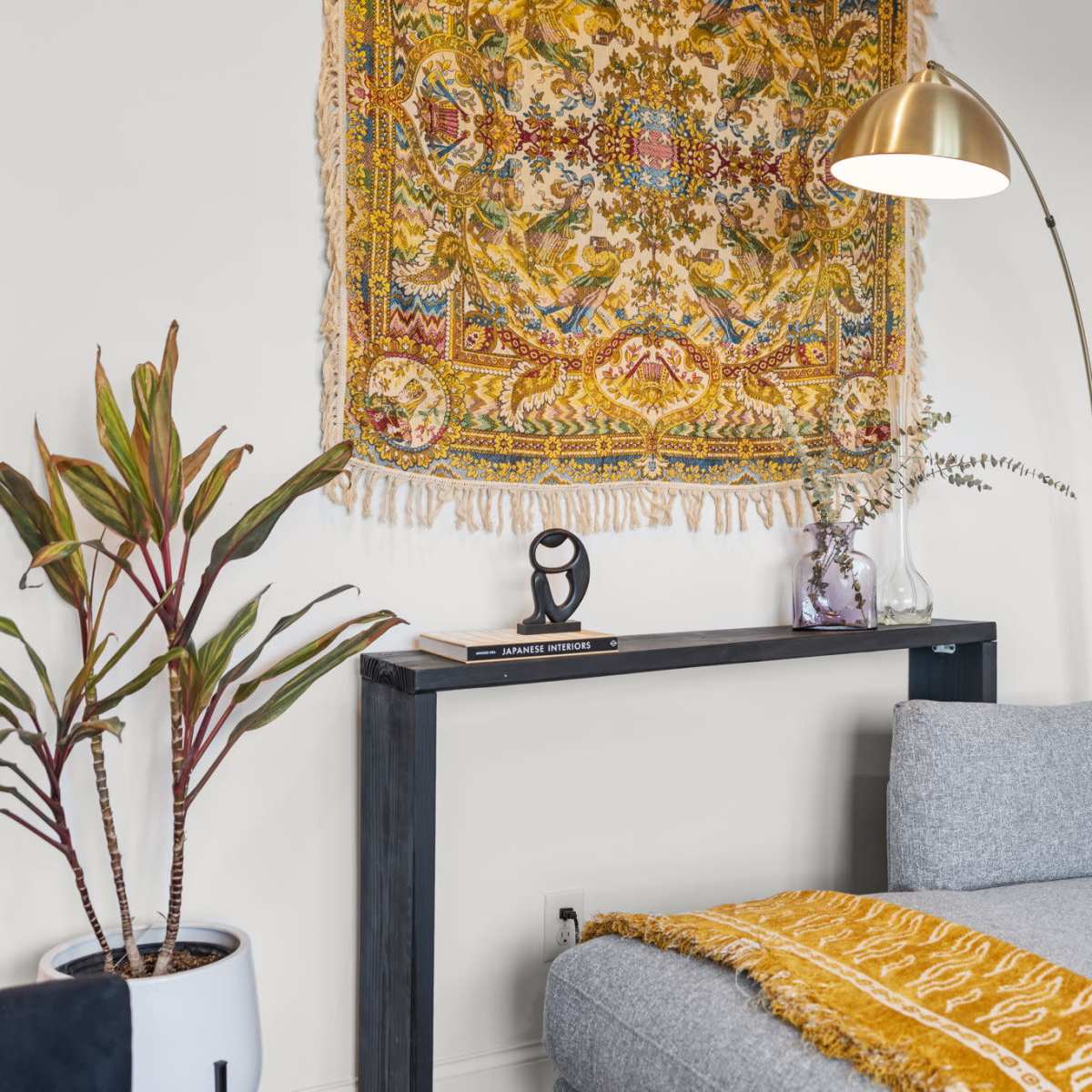 A cozy living space features a gray sectional sofa, a gold floor lamp, and a decorative wall tapestry above a console table with a plant and accessories.