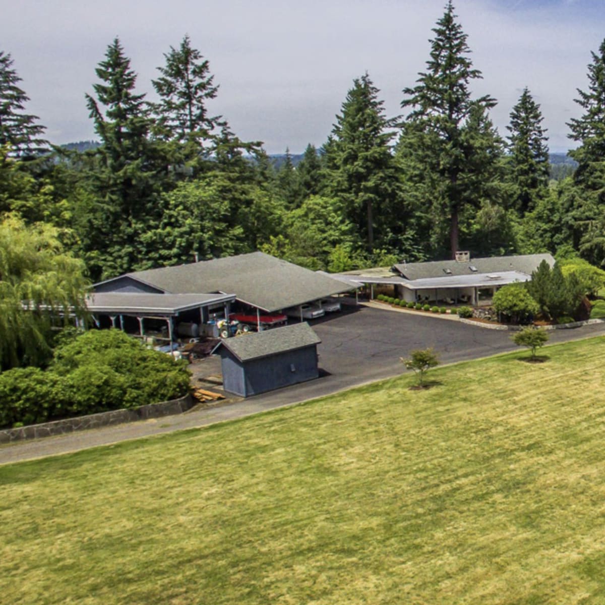 A sprawling house is situated on a grassy lawn surrounded by dense trees in a rural setting.