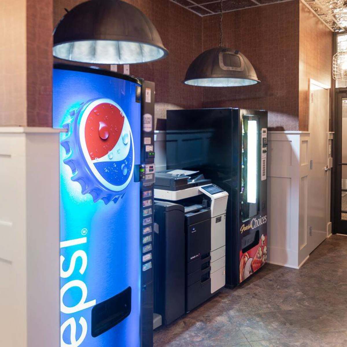 A vending area featuring a Pepsi vending machine, a printer, and a snack vending machine, with an adjacent glass door leading to another room.