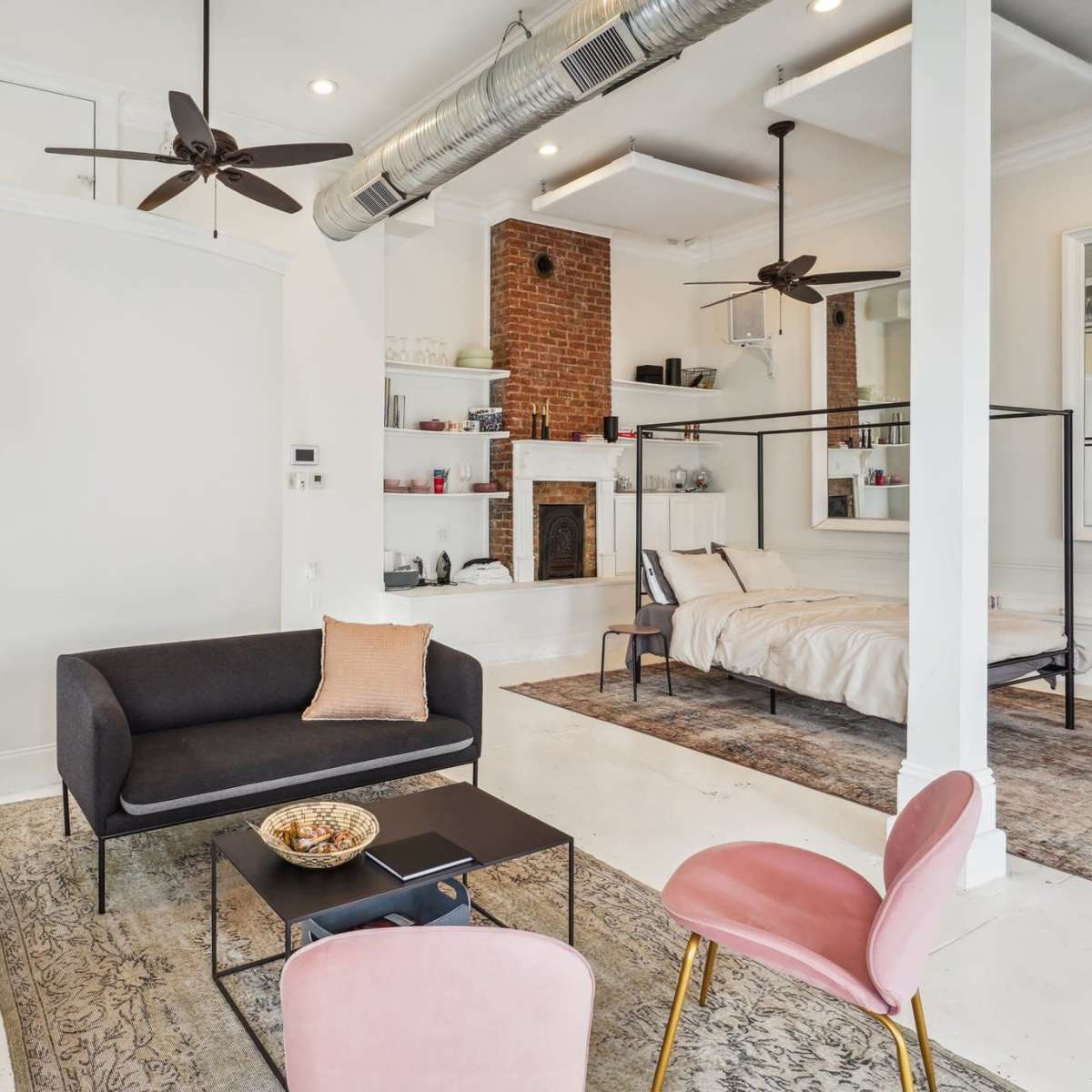A modern studio apartment featuring a living area with a sofa and television, a dining table, and a bedroom area with a four-poster bed, all set against a backdrop of exposed brick and sleek furnishings.