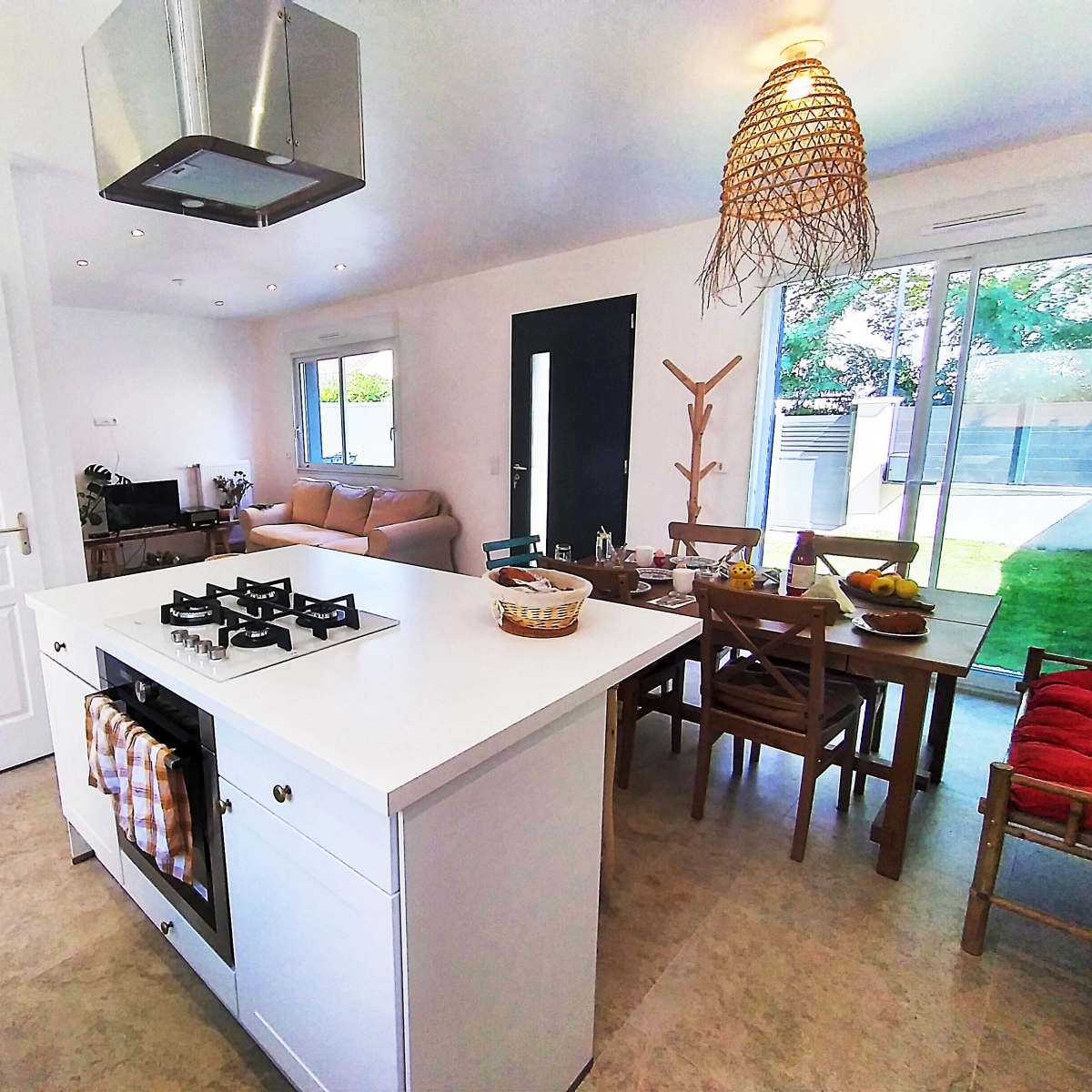 A modern kitchen with a central island, a gas stove, a dining area with wooden chairs, and large windows overlooking a garden.