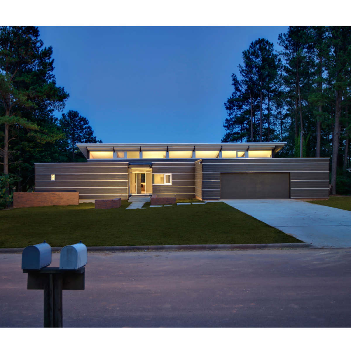 A modern, single-story house with horizontal siding and a large garage is set against a backdrop of tall trees, illuminated by exterior lighting at dusk.