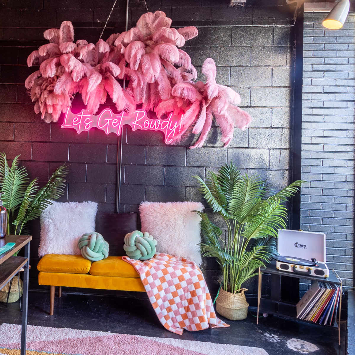 A cozy, stylish interior features a yellow couch with decorative pillows, a neon sign that says "Let's Get Rowdy," and plants arranged around a workspace with a computer and vinyl records.