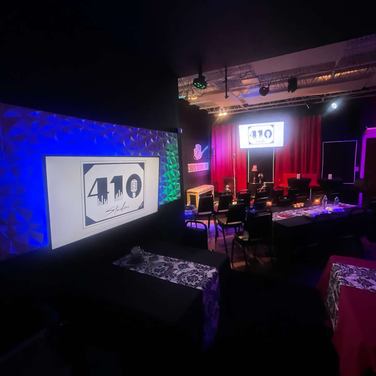 A dimly lit performance space features rows of black chairs and tables with colorful tablecloths, illuminated by various stage lights and a large banner displaying "410 Studio."
