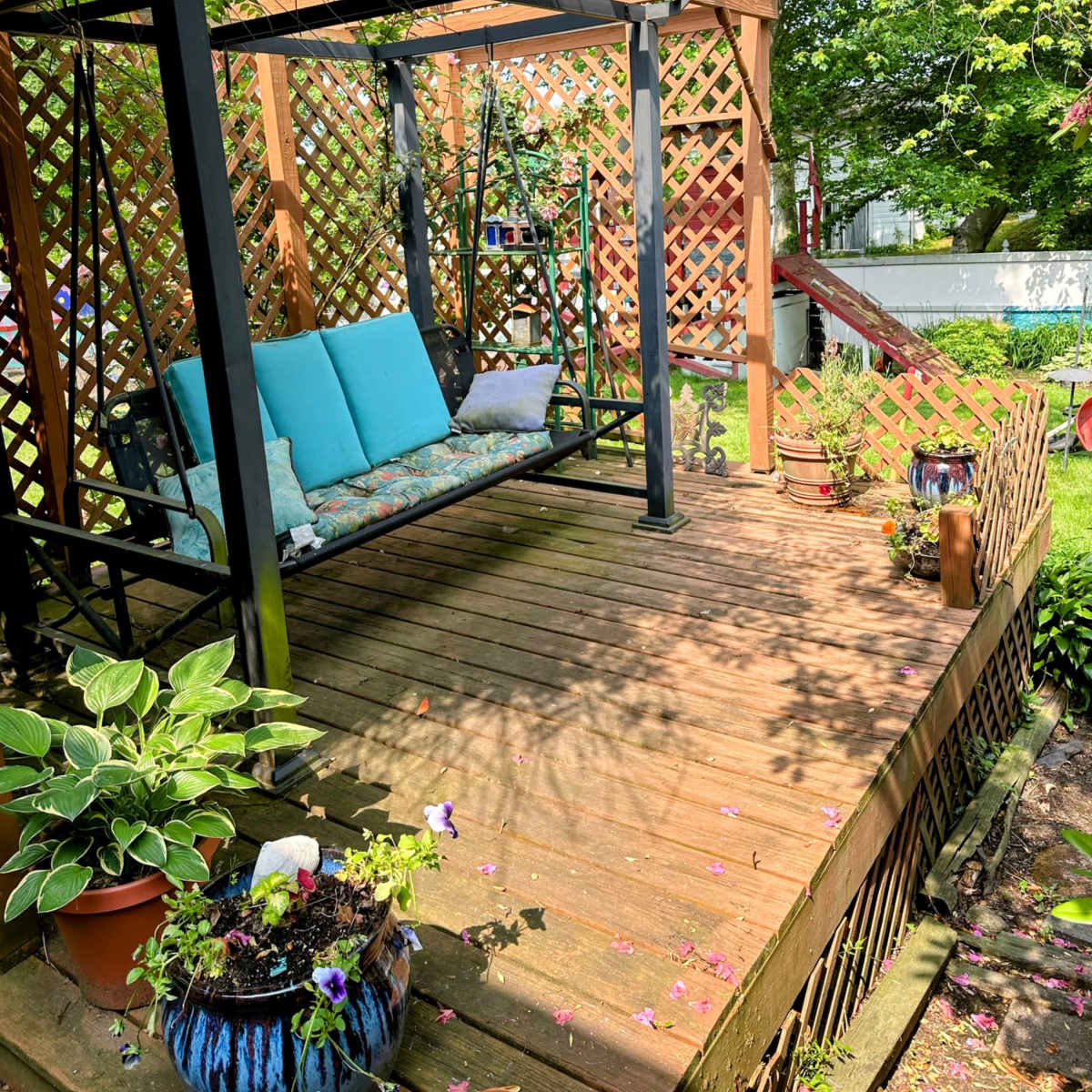 A wooden deck features a swing seat under a pergola with lattice panels, surrounded by potted plants and trees.