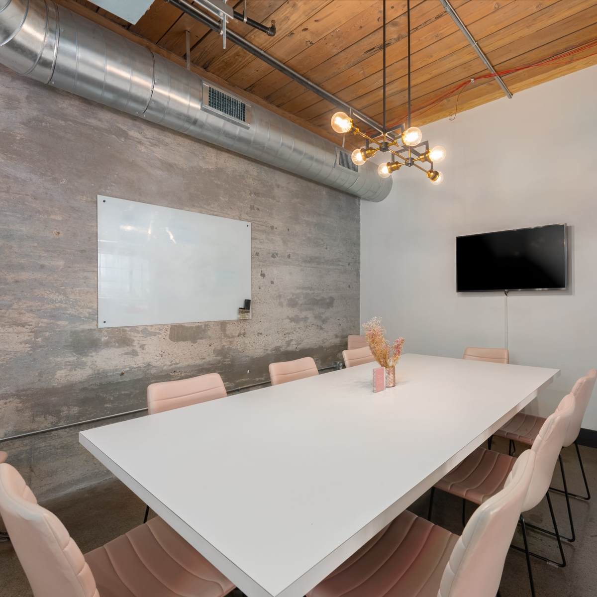 A modern conference room features a large white table surrounded by pink chairs, with a whiteboard and a TV mounted on the wall.