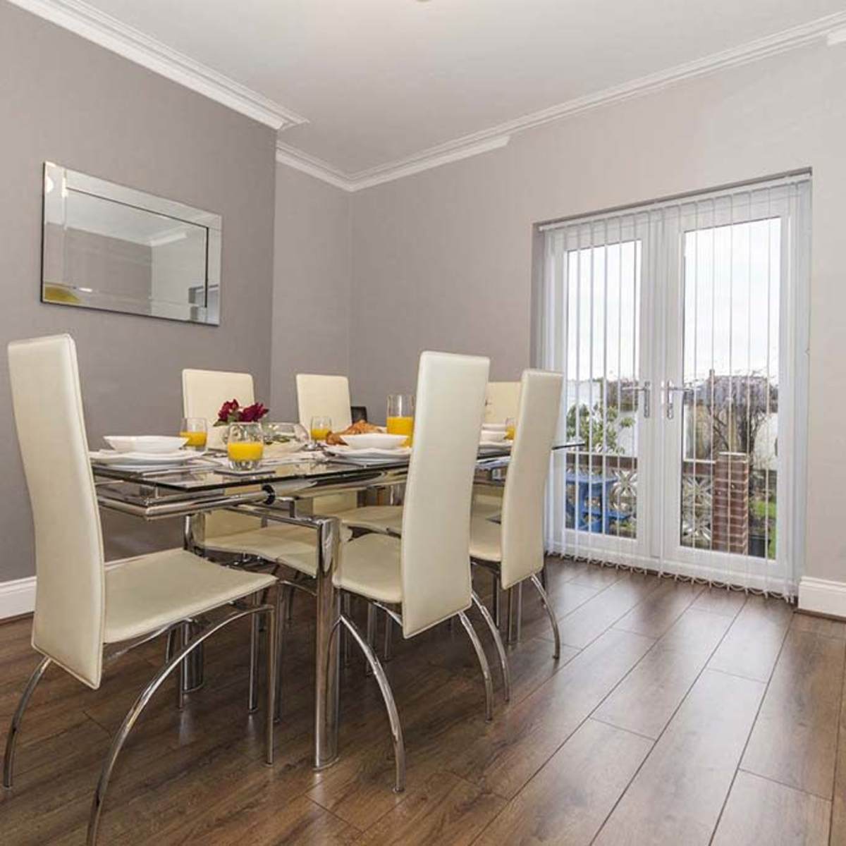 A modern dining area features a glass table with six light-colored chairs and a view through sliding glass doors.