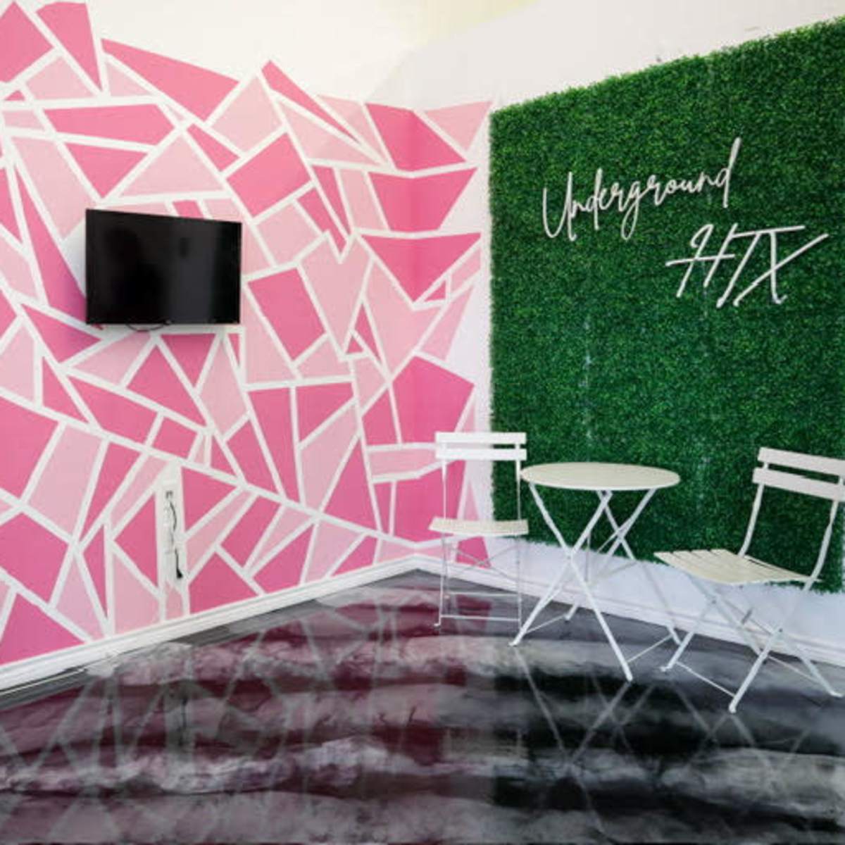 The room features pink geometric wall artwork, a green foliage accent wall with the text "Underground HTX," and a small white table with two chairs.