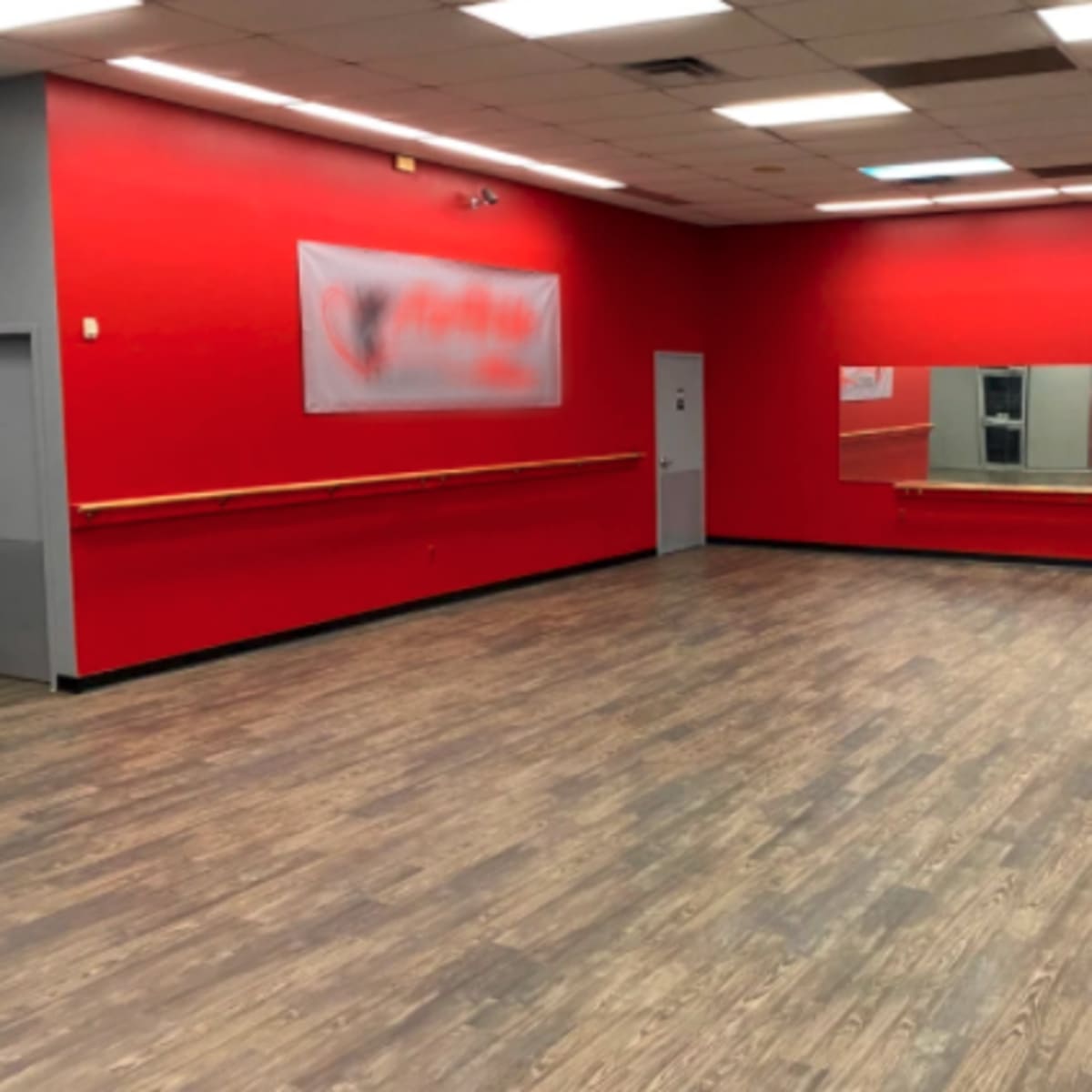An empty dance studio with red walls, a wooden floor, a long mirror on one side, and a barre along the wall.