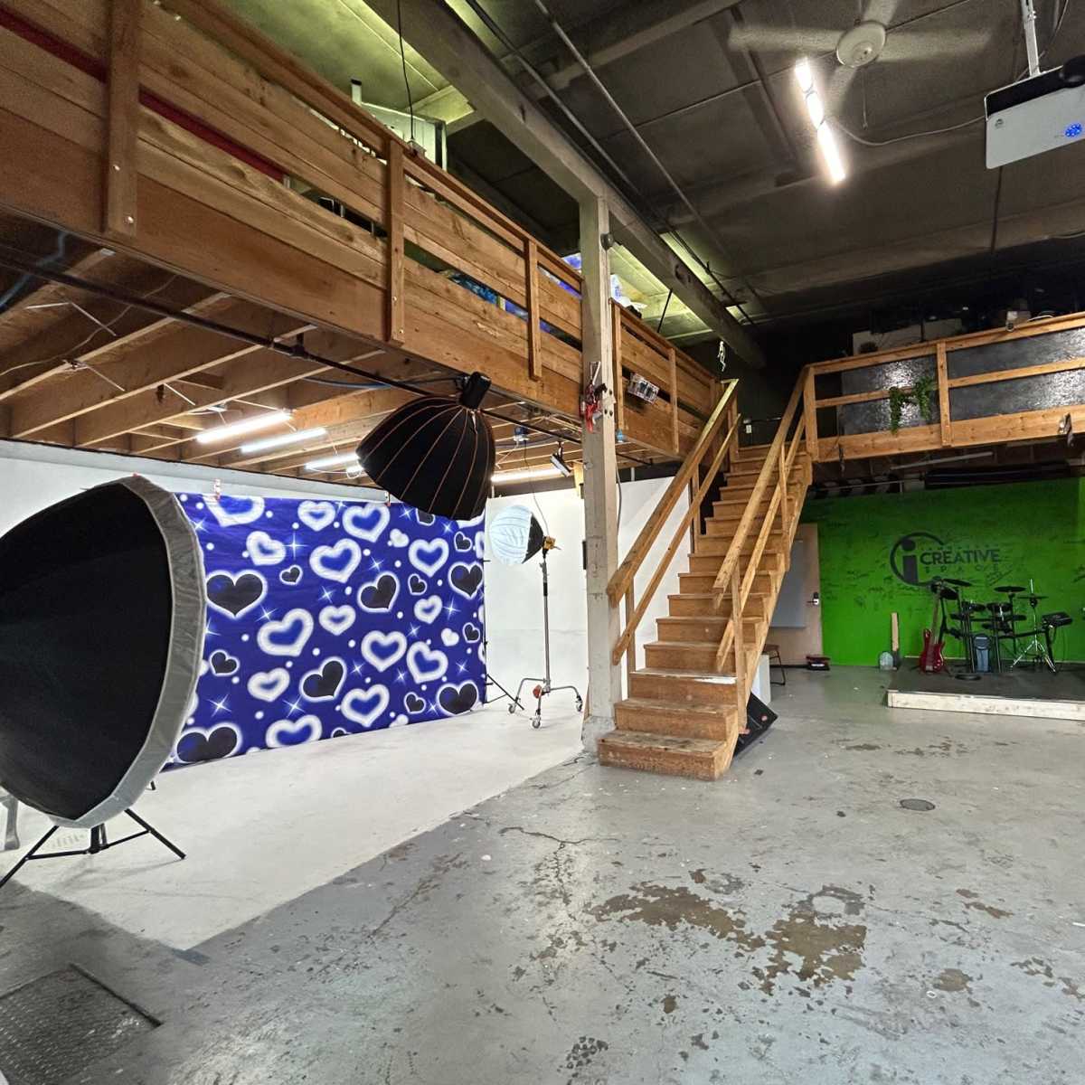 A spacious studio with a staircase leading to a loft area, equipped with photography backdrops and lighting equipment, as well as a green wall featuring the word "Creative."