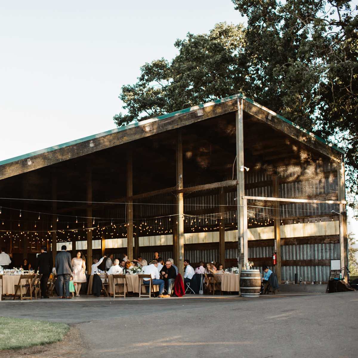 A large outdoor barn setting is filled with tables and guests celebrating, illuminated by string lights.