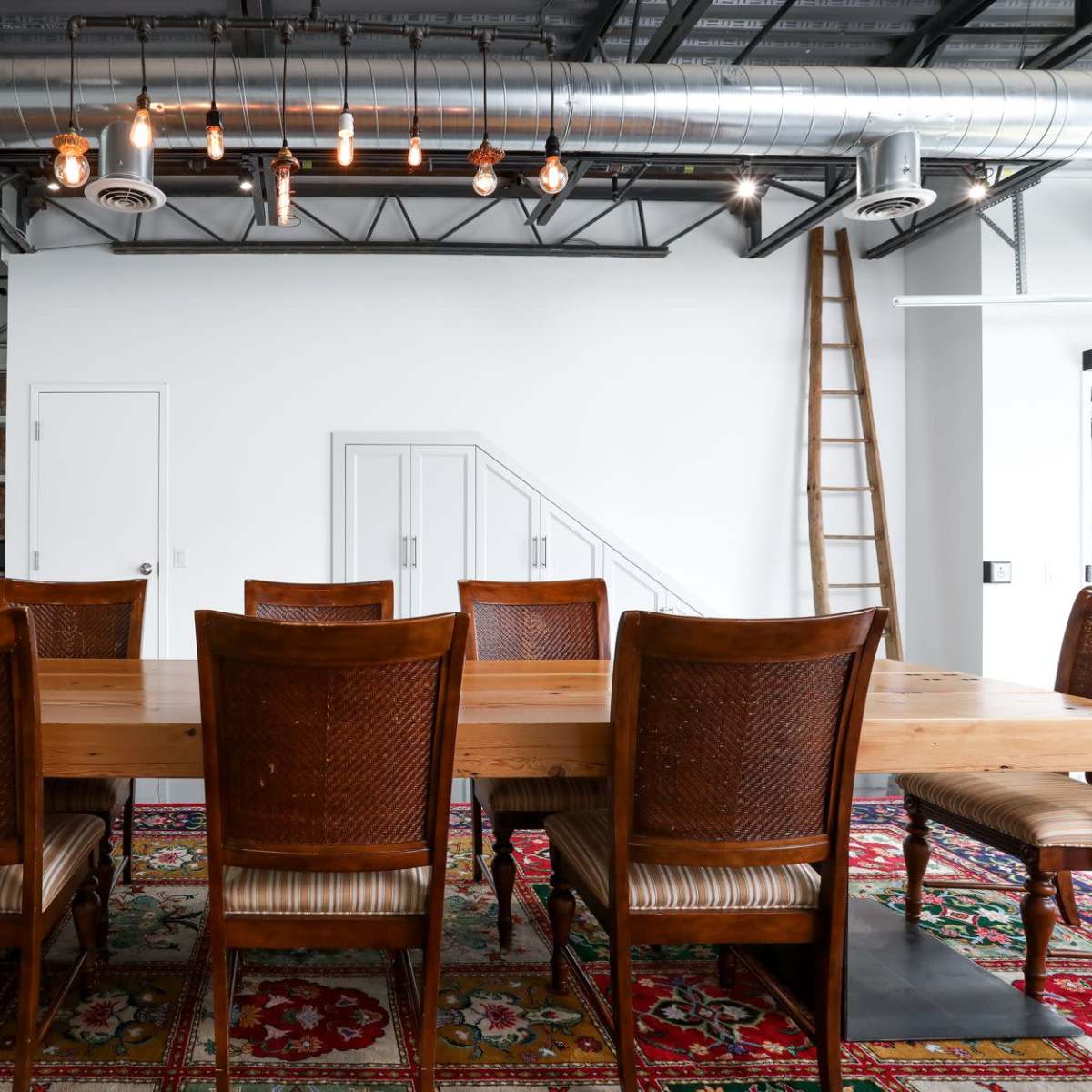 A long wooden dining table with eight brown chairs is set in a spacious room featuring exposed metal beams and soft lighting, with a patterned rug underneath.