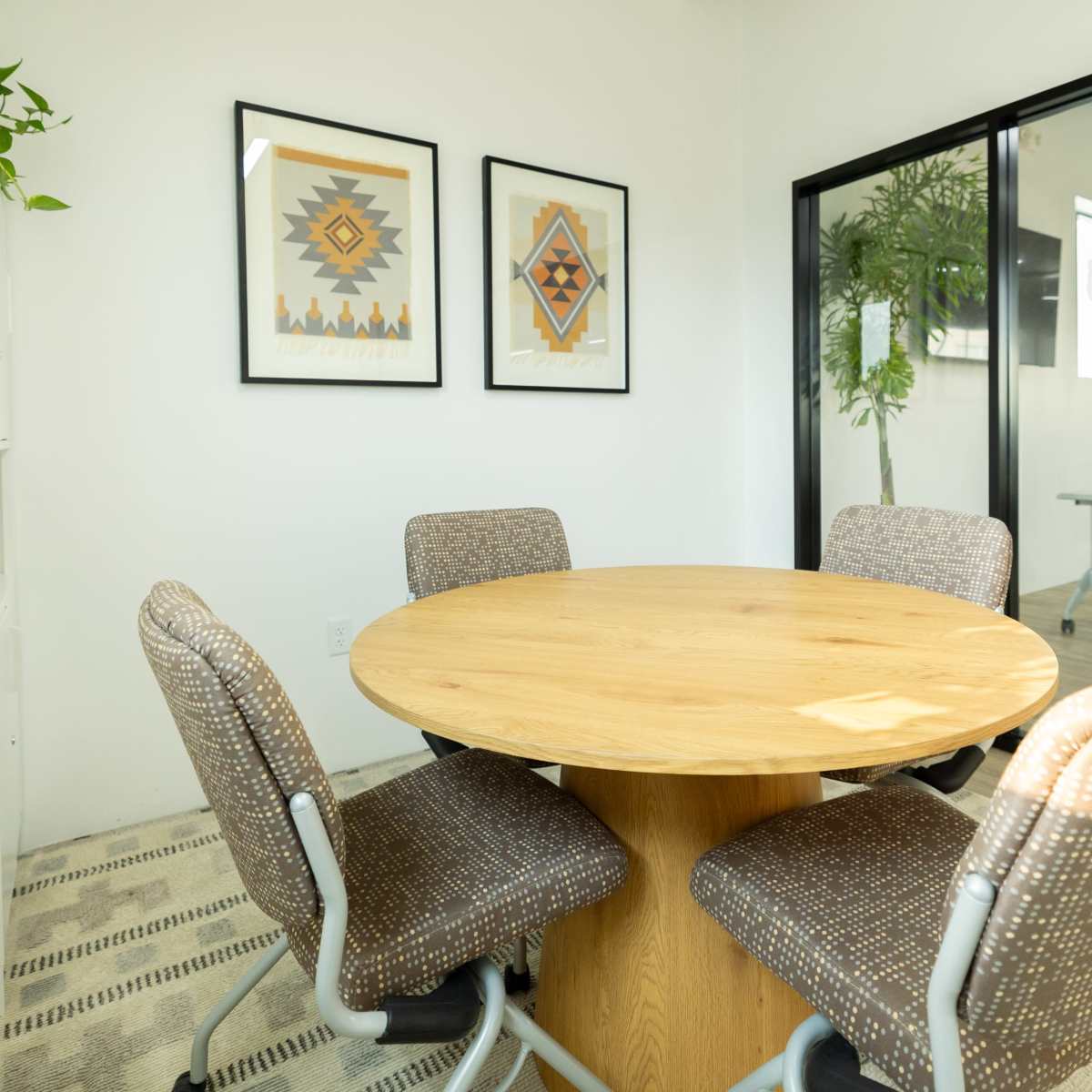 A small conference room featuring a round wooden table surrounded by four patterned chairs, with framed artwork on the walls and glass partitions.