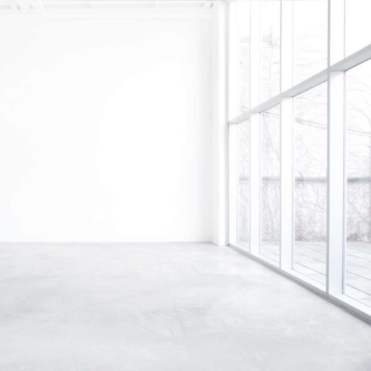 A minimalistic, empty room with a gray concrete floor and large windows letting in natural light.