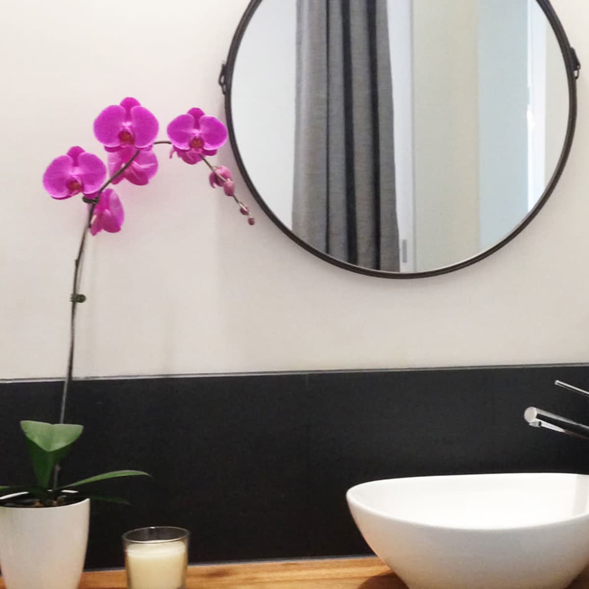 A modern bathroom with a round mirror, a sink, a potted orchid, and a candle on a wooden countertop.