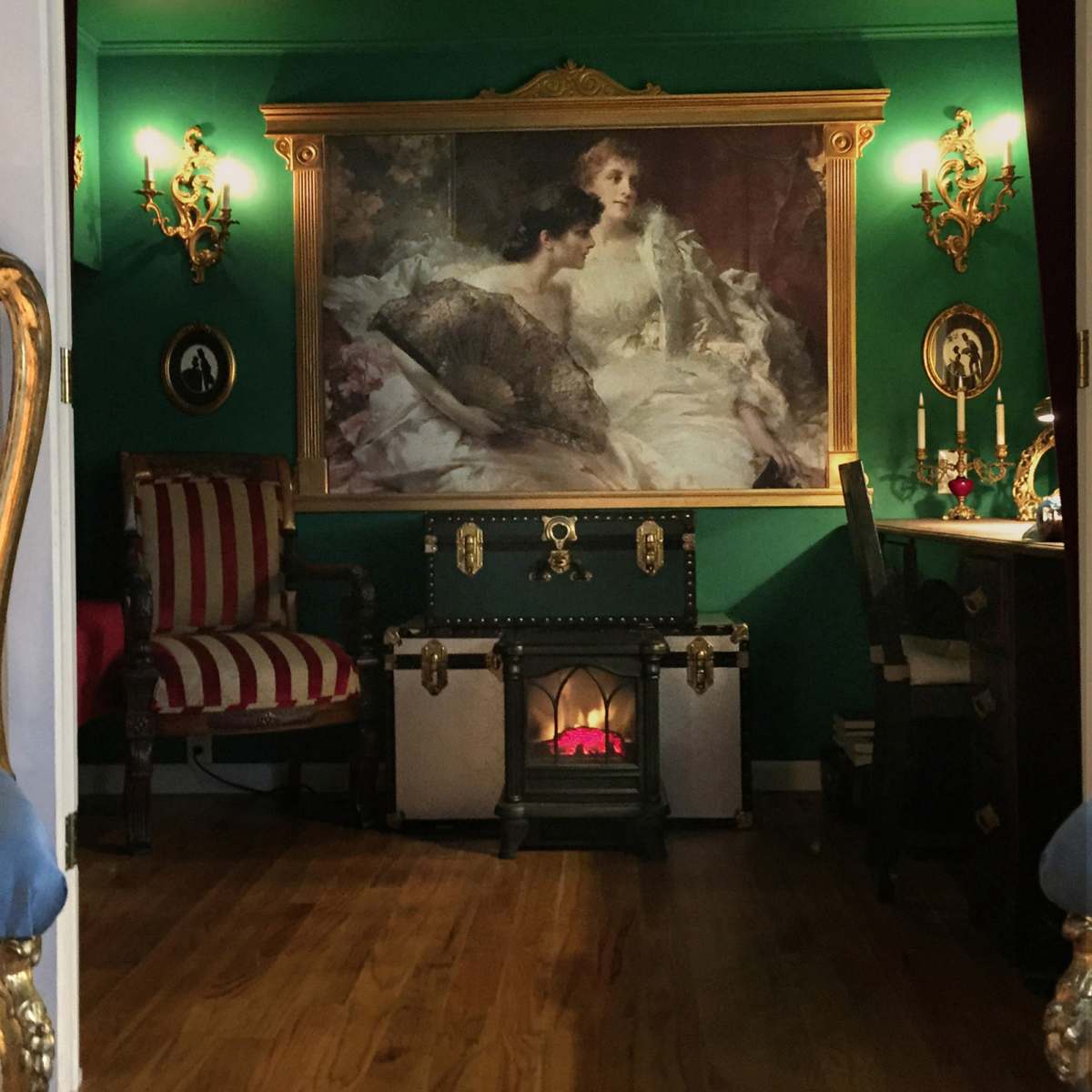 A room with ornate golden chairs facing a large painting on a green wall, flanked by decorative sconces and a fireplace.