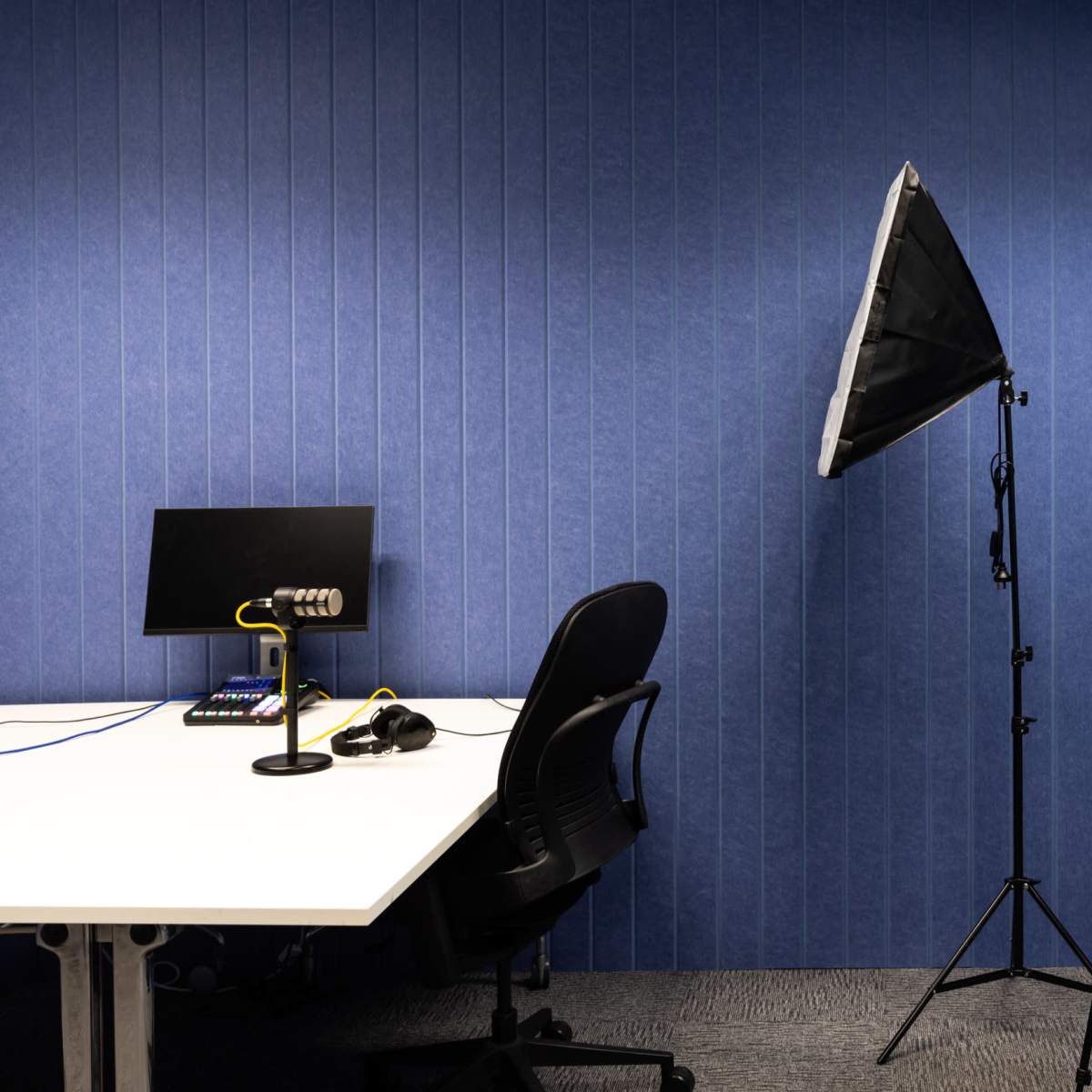A small recording studio with a desk equipped with a computer, a microphone, and a chair, alongside lighting equipment and a backdrop.