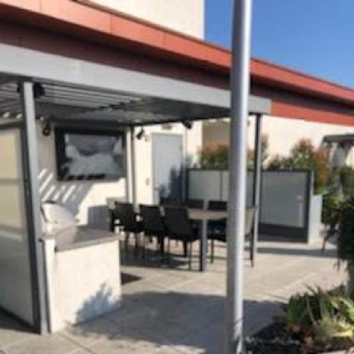A patio area with a dining table and chairs, surrounded by walls and a grill, under a pergola on a clear day.