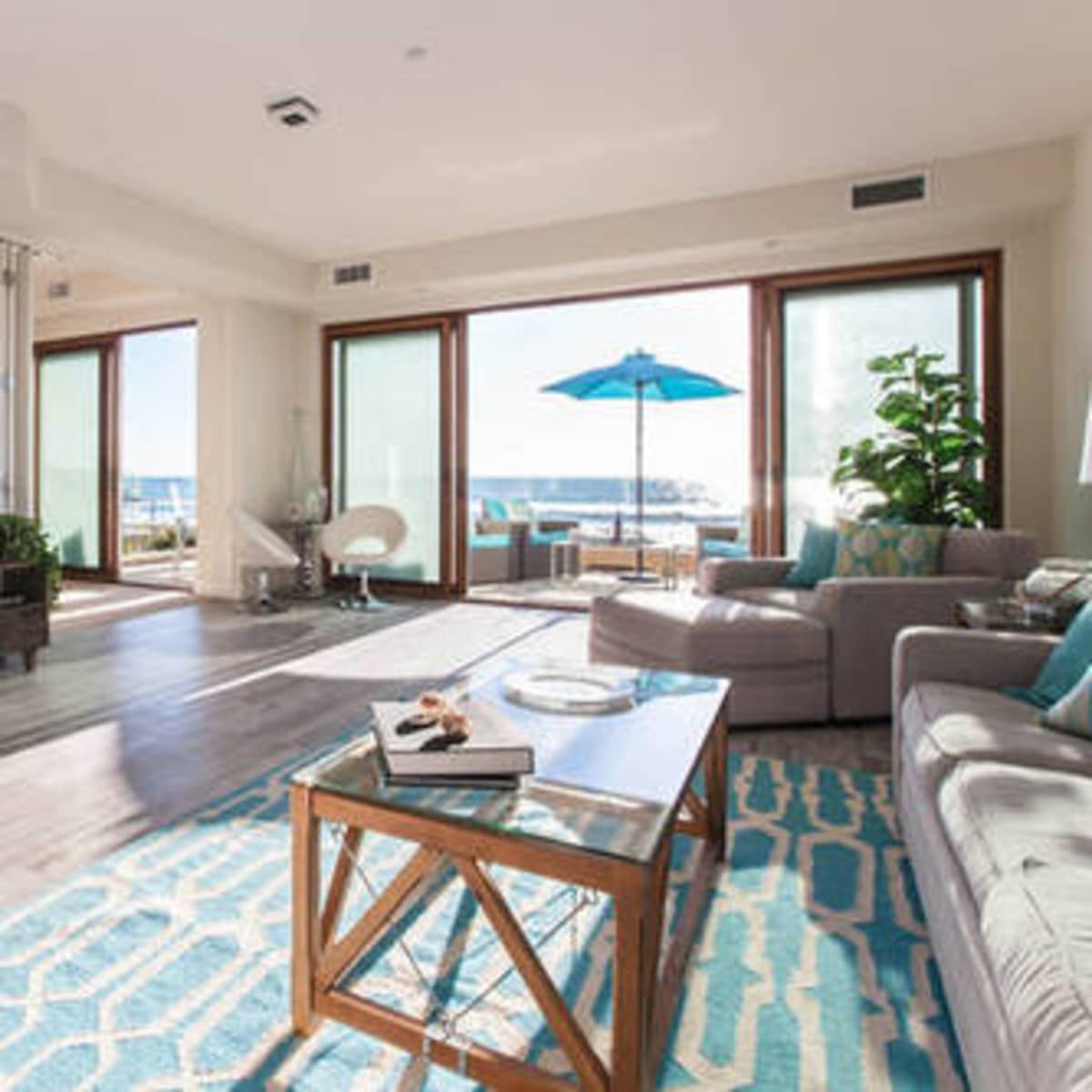 A bright, modern living room with large windows overlooking a patio featuring an umbrella, a comfortable seating area, and a fireplace.