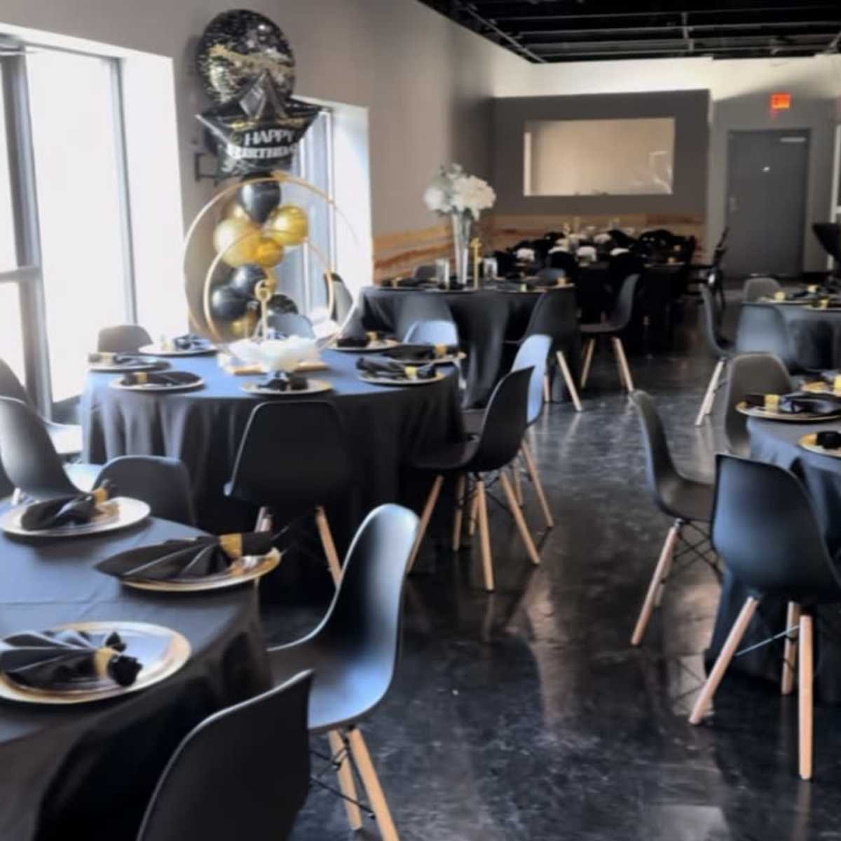 A decorated event space with multiple tables set for a celebration, featuring black tablecloths, gold and black balloons, and floral centerpieces.