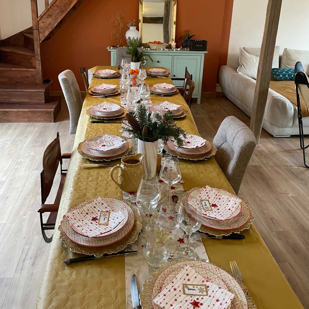A dining table is set with patterned plates and glassware in a warmly lit room featuring a staircase and an orange accent wall.