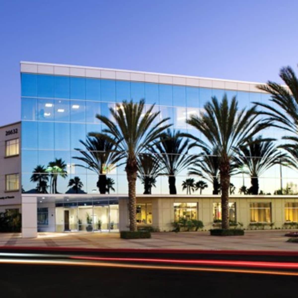 A modern, multi-story building with large glass windows reflecting palm trees and a street in the foreground at dusk.