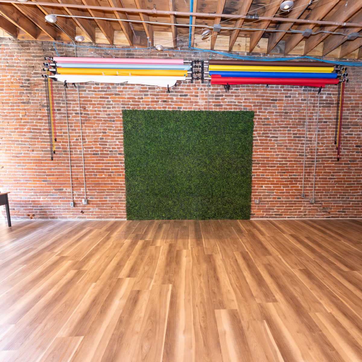 A spacious, empty studio featuring a brick wall with a green grass backdrop and colorful backdrops mounted above, alongside a wooden floor.