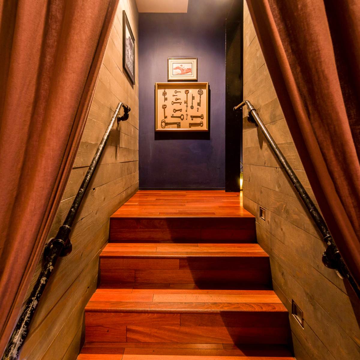 A staircase leads upward through a pair of curtains, with a framed display of keys on the wall at the top.