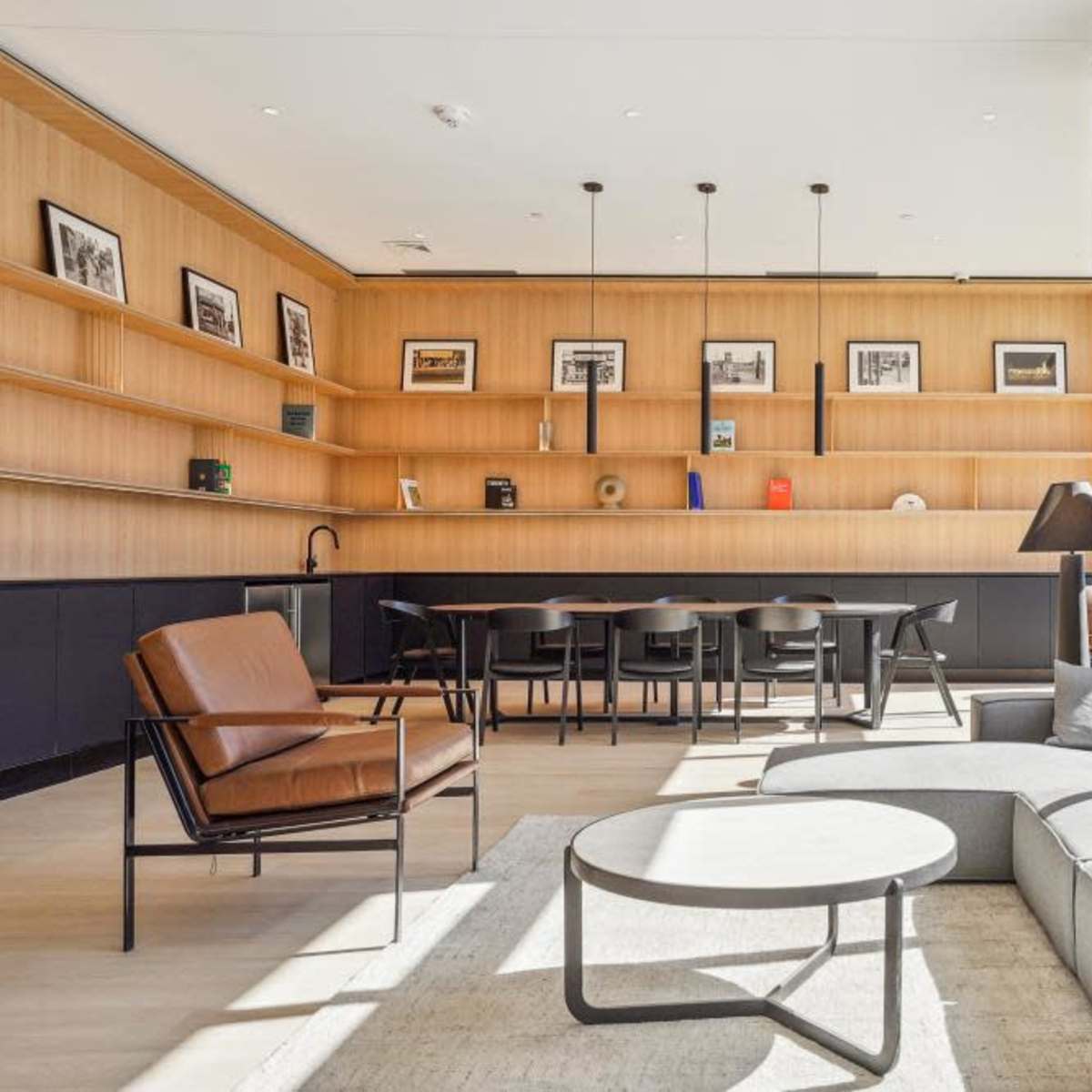 A modern interior space featuring a cozy seating area with a curved gray sofa and a brown armchair, along with a wooden shelf displaying framed pictures and a dining area in the background.