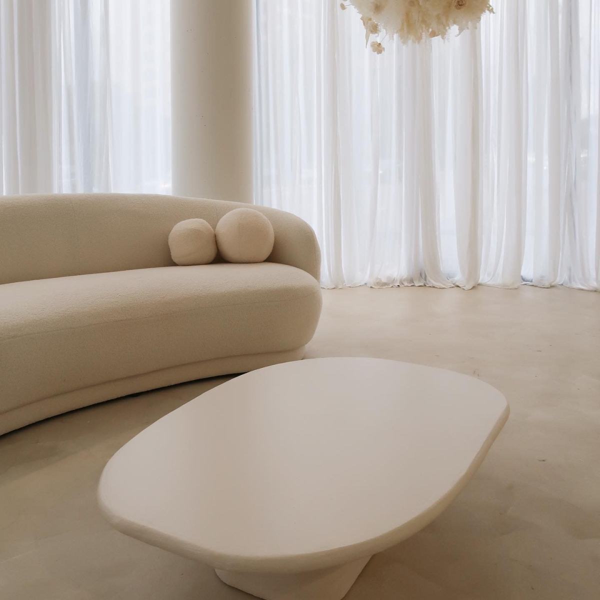 A curved, light-colored sofa with two round cushions is positioned near a sculptural, white coffee table in a bright, airy room with sheer curtains.