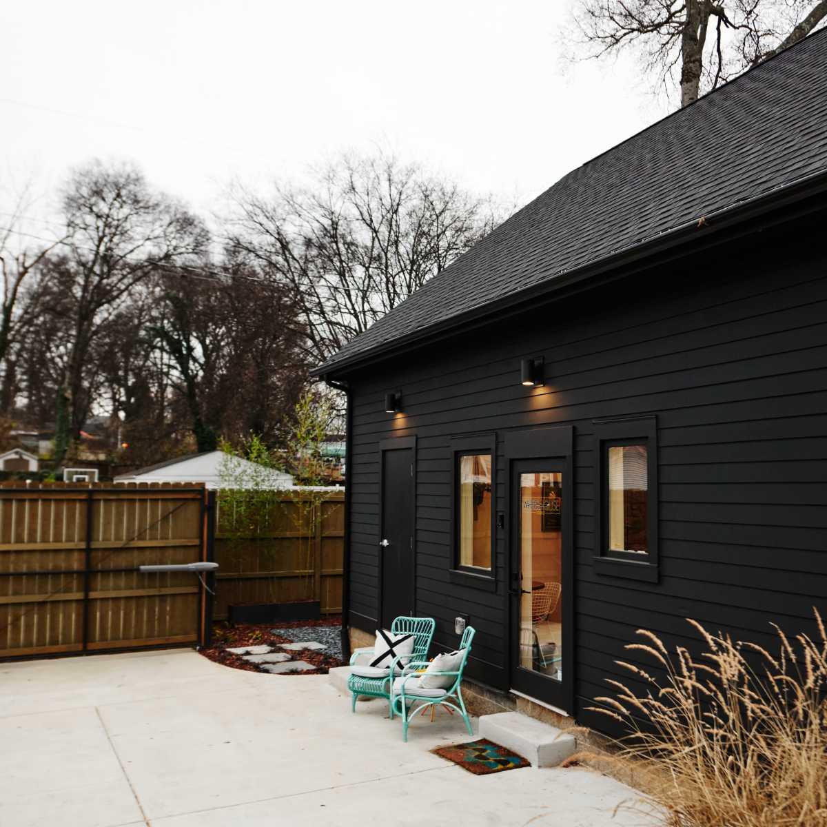 A black wood-sided house features a small patio area with a couple of light blue chairs and a fenced yard in the background.