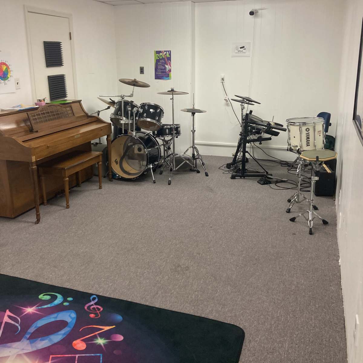 A music practice room featuring an acoustic piano on the left, a drum set in the center, and an electronic drum kit against the wall.