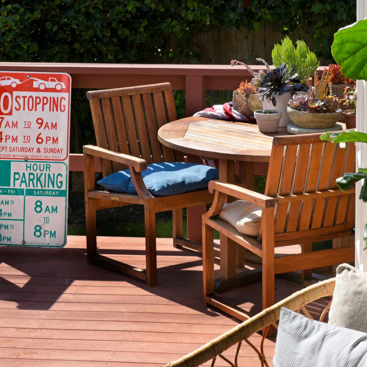 A wooden patio area with a round table and two chairs, decorated with potted plants and cushions, alongside a parking sign.
