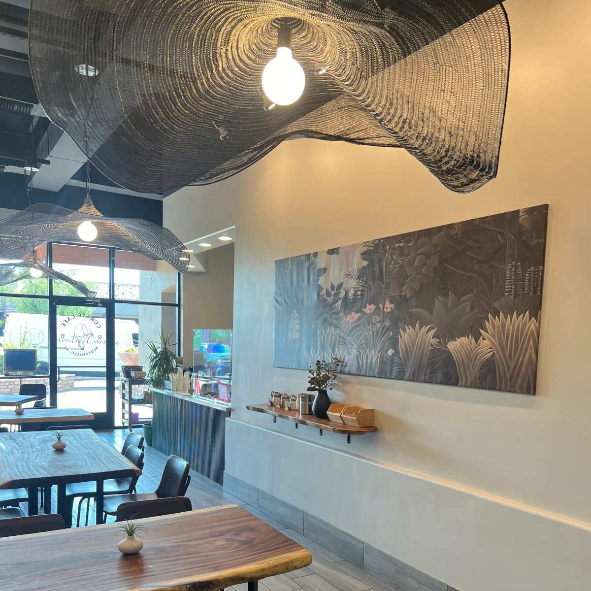 A modern cafe interior with wooden tables, black chairs, and a wall adorned with a large artwork surrounded by decorative hanging elements.