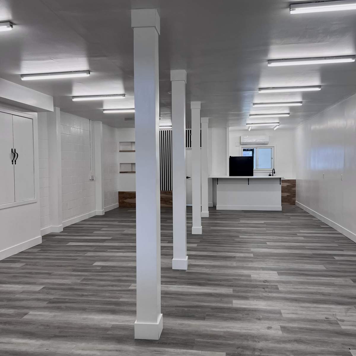 An empty room with gray wooden flooring, white walls, and several white supporting columns, featuring a kitchen area in the back.