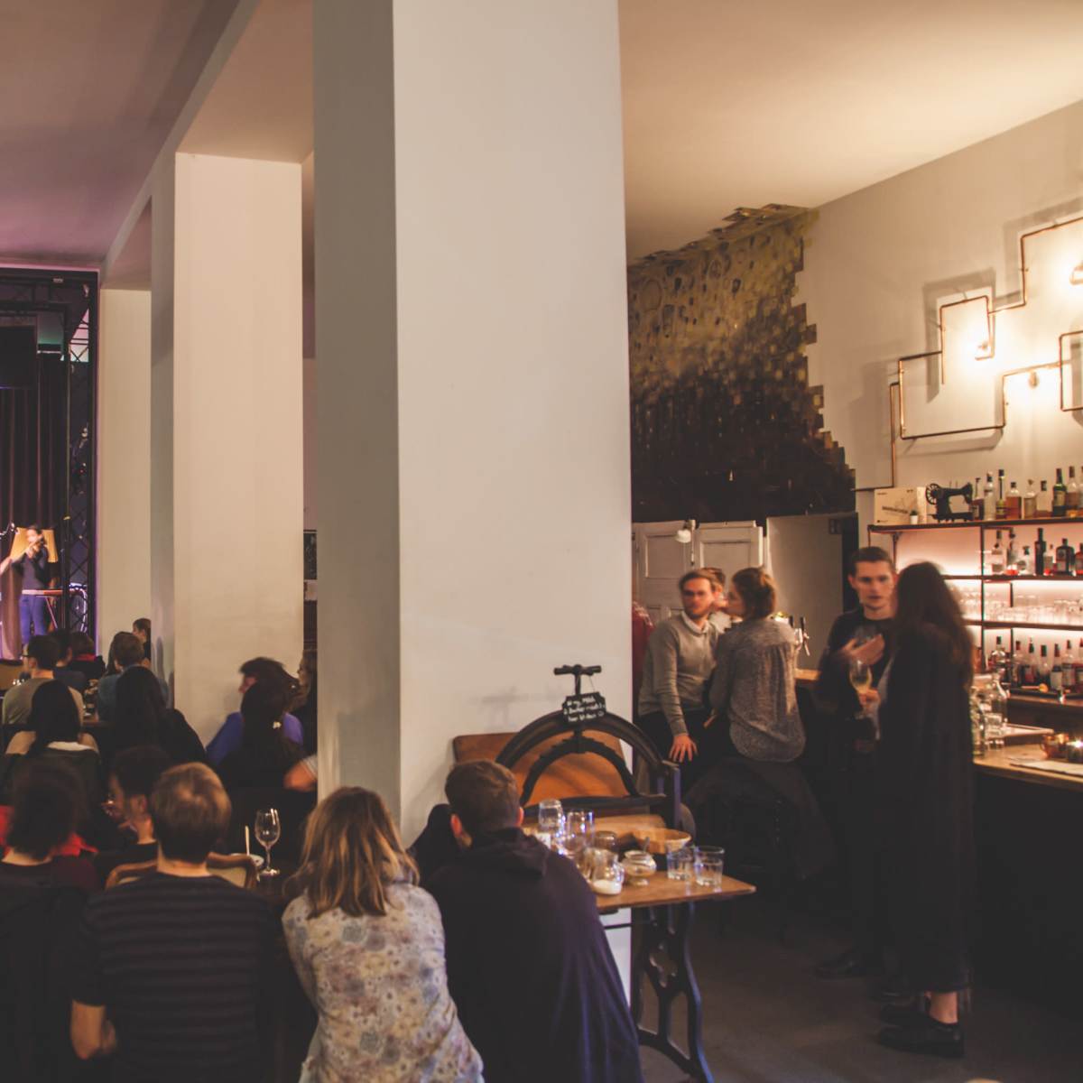 A performer plays guitar on stage in a dimly lit venue filled with an audience seated at tables and standing around the bar.