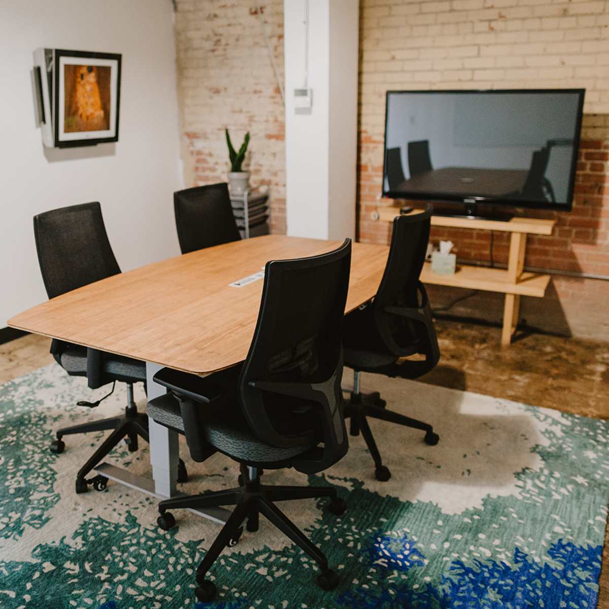 A conference room features a large wooden table surrounded by five black rolling chairs, with a television mounted on the wall and a patterned rug on the floor.