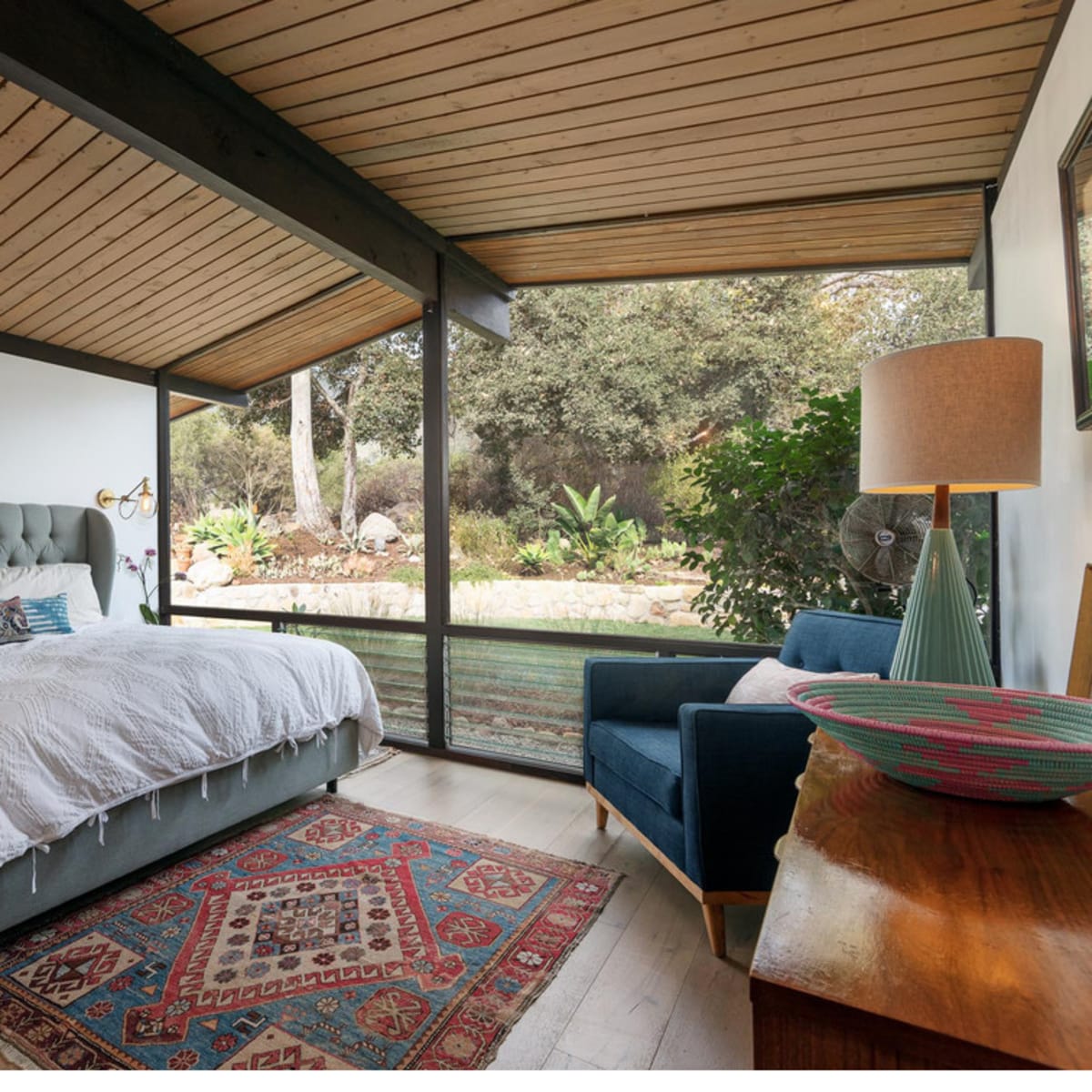 A modern bedroom features a tufted bed, a blue armchair, and large glass windows overlooking a garden.