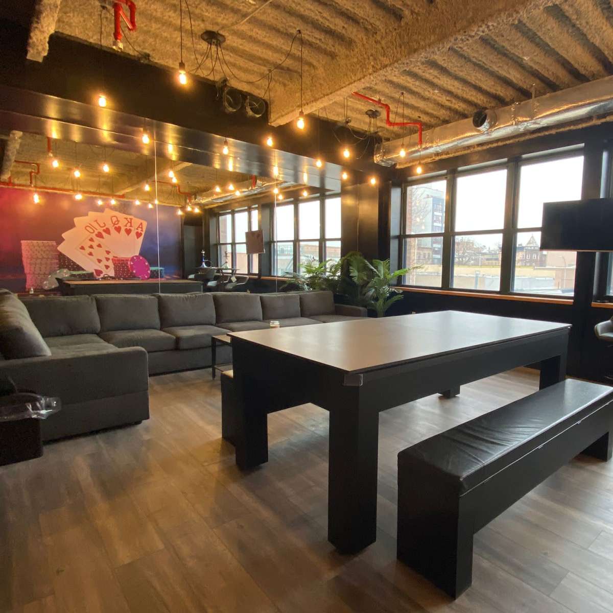 A modern office space featuring a large black table with a bench, surrounded by comfortable seating and large windows allowing natural light to fill the room.